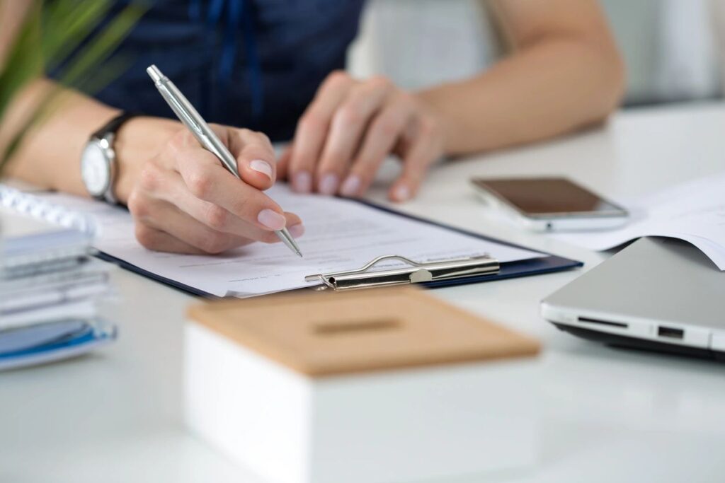 A person is writing on a clipboard at a desk with a laptop and phone nearby.