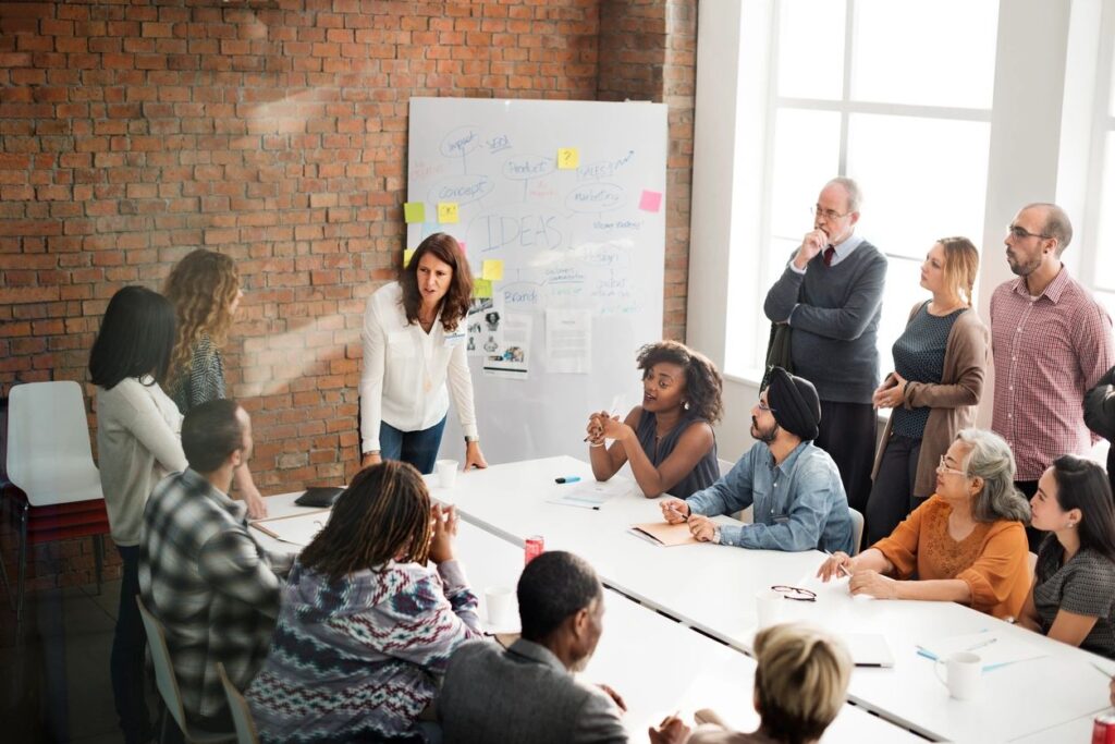A diverse group in a meeting, discussing ideas with a presenter at a whiteboard covered in notes and stickies.