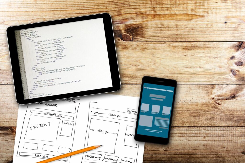 An iPad displays code while a smartphone shows a digital design, alongside a sketch of a website layout and a pencil.